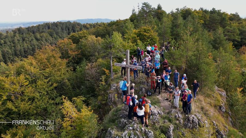 Menschen stehen auf einem bewaldeten Berg mit Gipfelkreuz