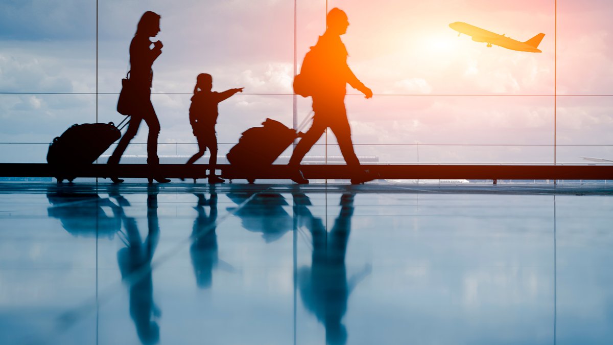 Silhouette einer jungen Familie mit Koffern, im Hintergrund ein startendes Flugzeug