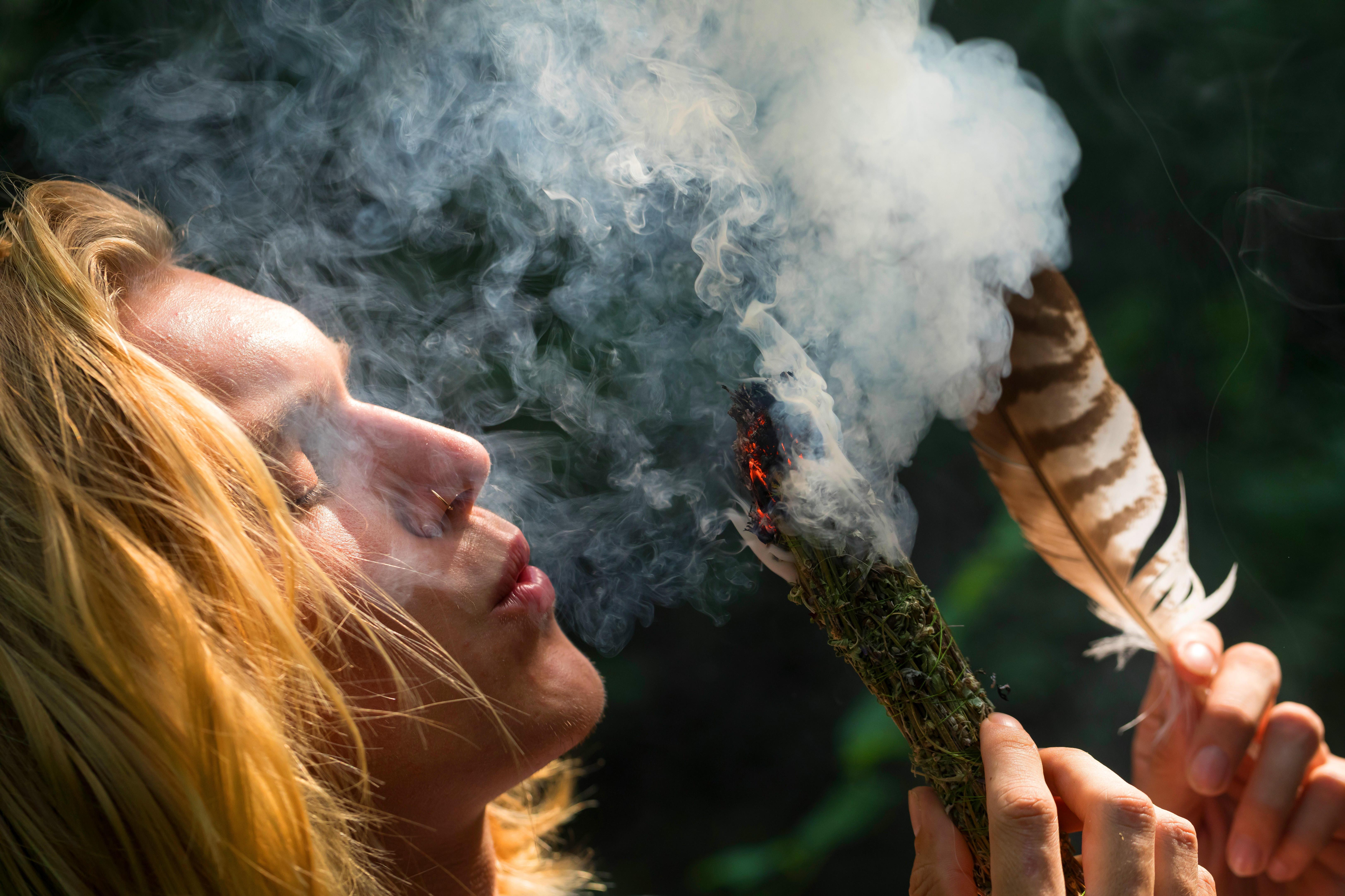 Eine Person pustet in Richtung eines glimmenden Kräuterbündels und hat eine Feder in der Hand.