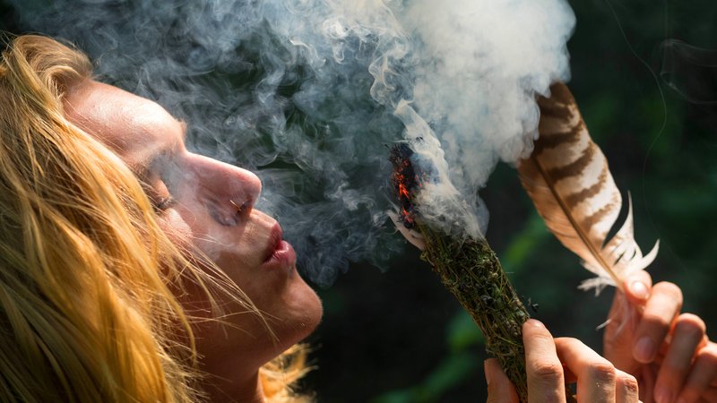 Eine Person pustet in Richtung eines glimmenden Kräuterbündels und hat eine Feder in der Hand. | Bild: picture alliance / blickwinkel/F. Hecker | F. Hecker Eine Person pustet in Richtung eines glimmenden Kräuterbündels und hat eine Feder in der Hand.