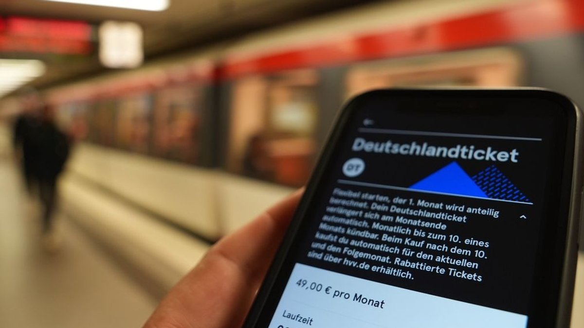 Ein Fahrgast hält ein Smartphone mit einem digitalen Deutschlandticket an einer U-Bahnstation in der Hand.