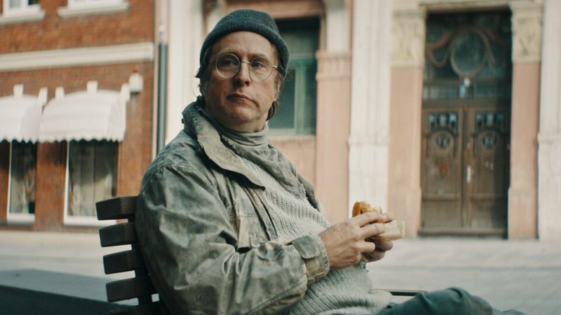 Ein Mann mit Mütze sitzt auf einem Stuhl auf der Straße und schaut ein wenig provozierend in die Kamera:Bjarne Mädel in "Faking Bullshit" (Filmszene)