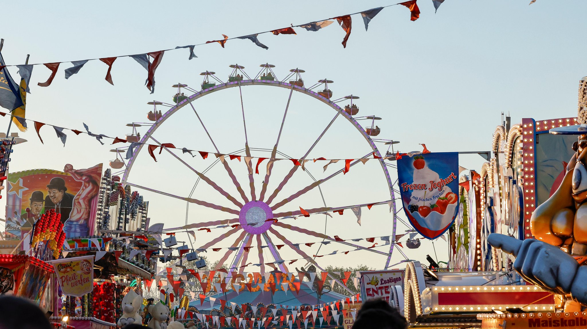 Ein Riesenrad dreht sich im Hintergrund, davor Wimpelgirlanden, Jahrmarktsbuden, Lichter: Volksfestkulisse