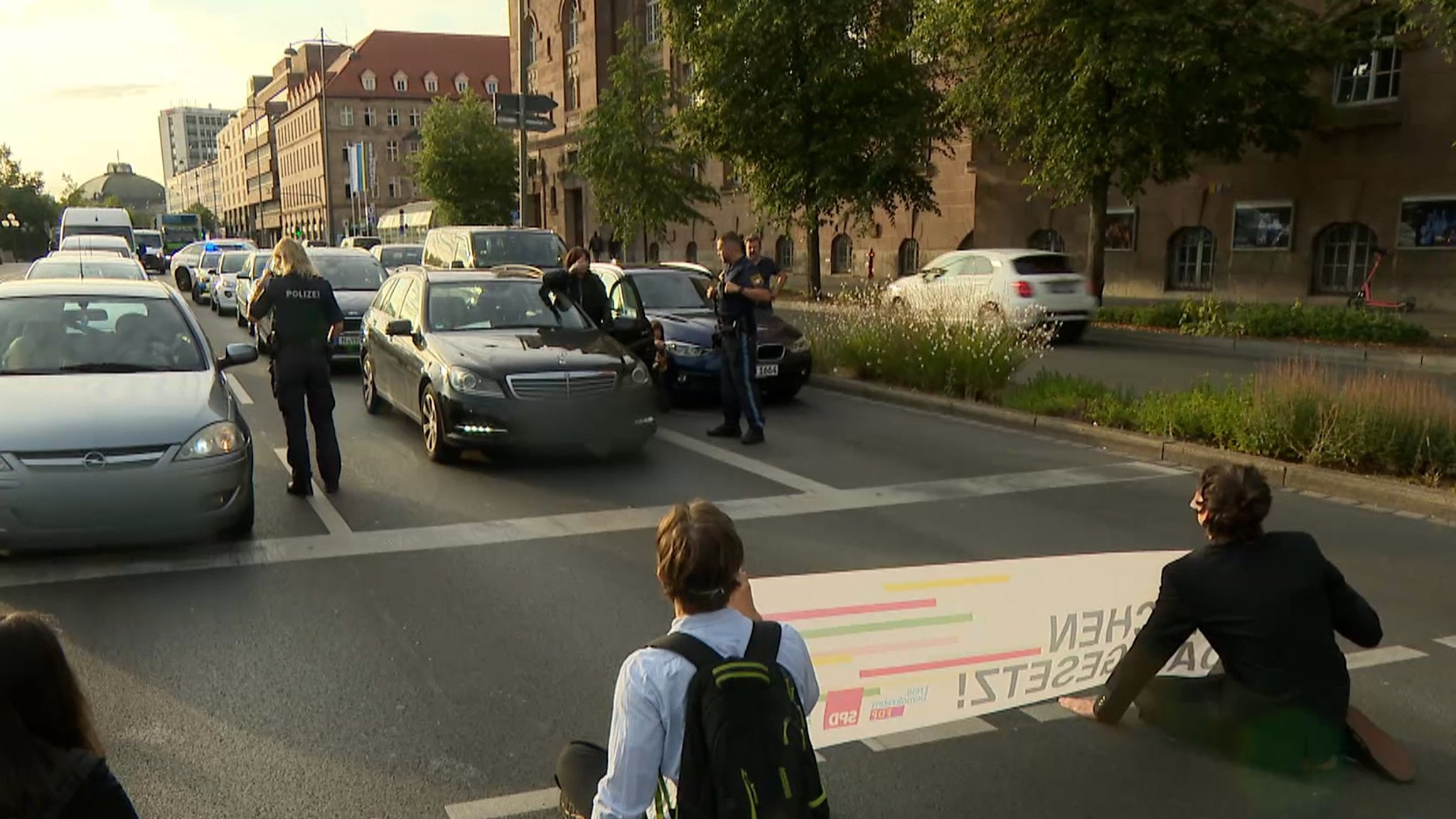 Menschen kleben auf de Straße, mit Polizei und wartenden Autofahrern.