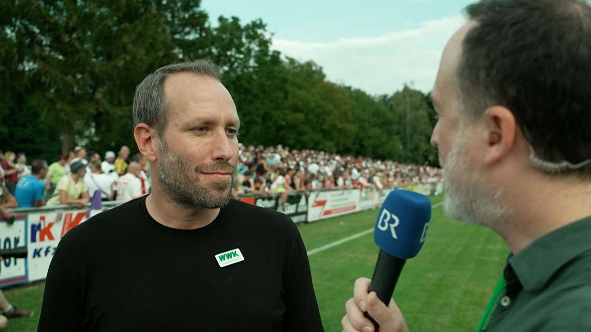 BR-Reporter Thomas Pösl steht während des Benefizspiels des FC Augsburg gegen den SSV Ulm am Spielfeldrand und interviewt FCA-Geschäftsführer Michael Ströll.