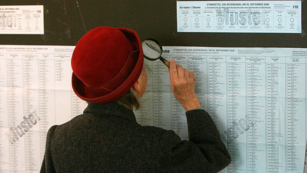Eine Frau liest mit einer Lupe am Wahlsonntag in Wolfratshausen die ausgehängten Musterstimmzettel. 
