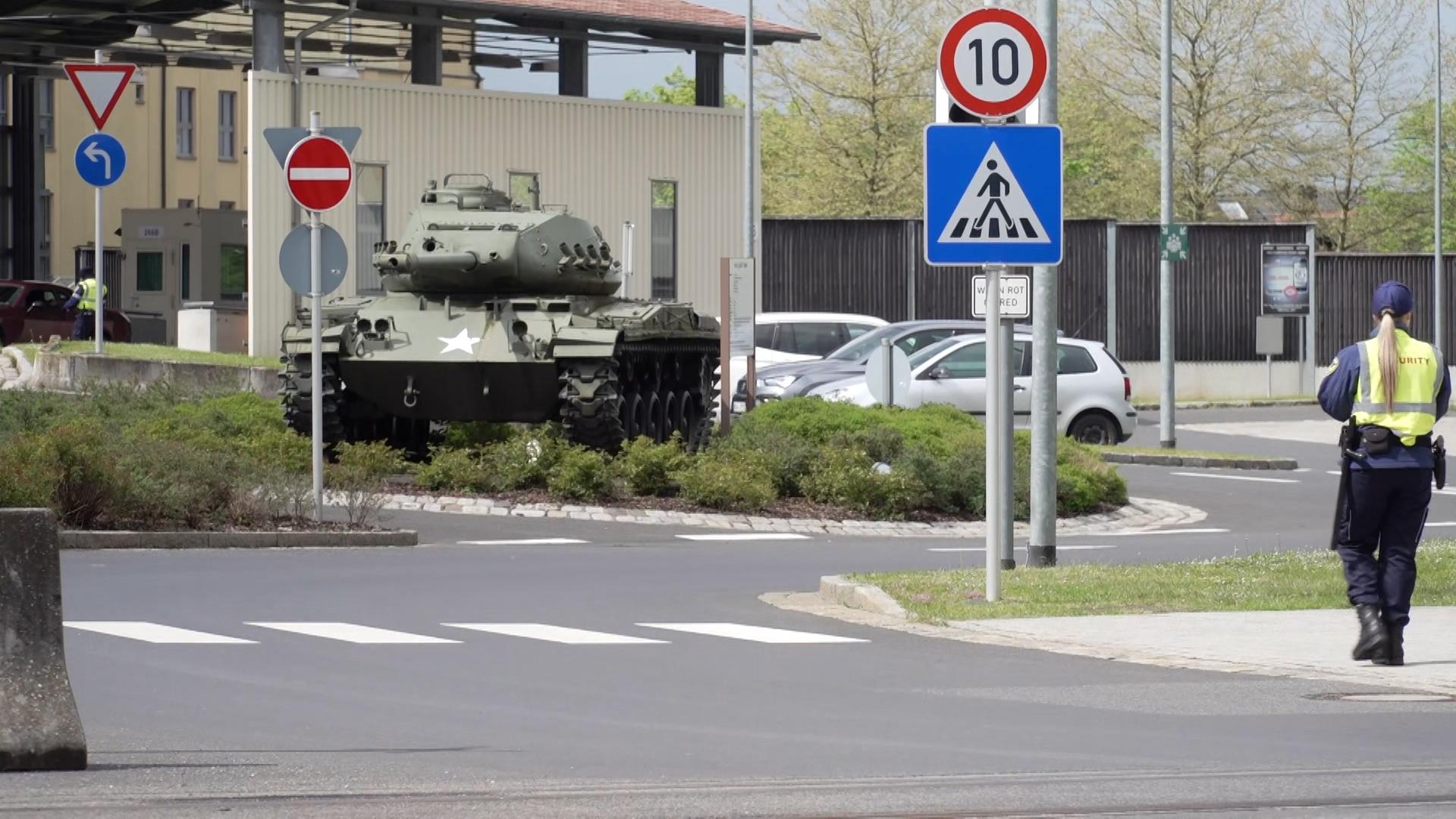 Ein Blick auf das Gelände der US-Armee in Grafenwöhr. Zu sehen ist ein Panzer, mehrere geparkte Autos und eine Frau, die eine gelbe Securityweste und Ausrüstung wie eine Pistole und einen Schlagstock bei sich trägt. 