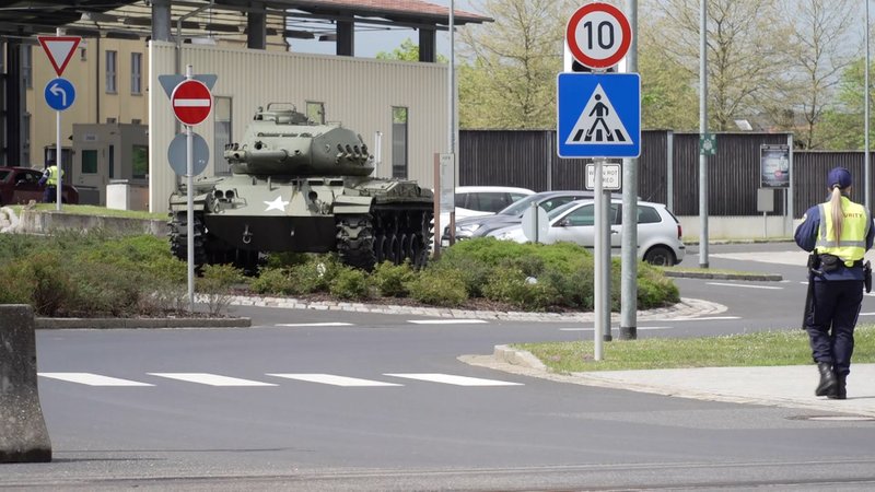 Ein Blick auf das Gelände der US-Armee in Grafenwöhr. Zu sehen ist ein Panzer, mehrere geparkte Autos und eine Frau, die eine gelbe Securityweste und Ausrüstung wie eine Pistole und einen Schlagstock bei sich trägt. 