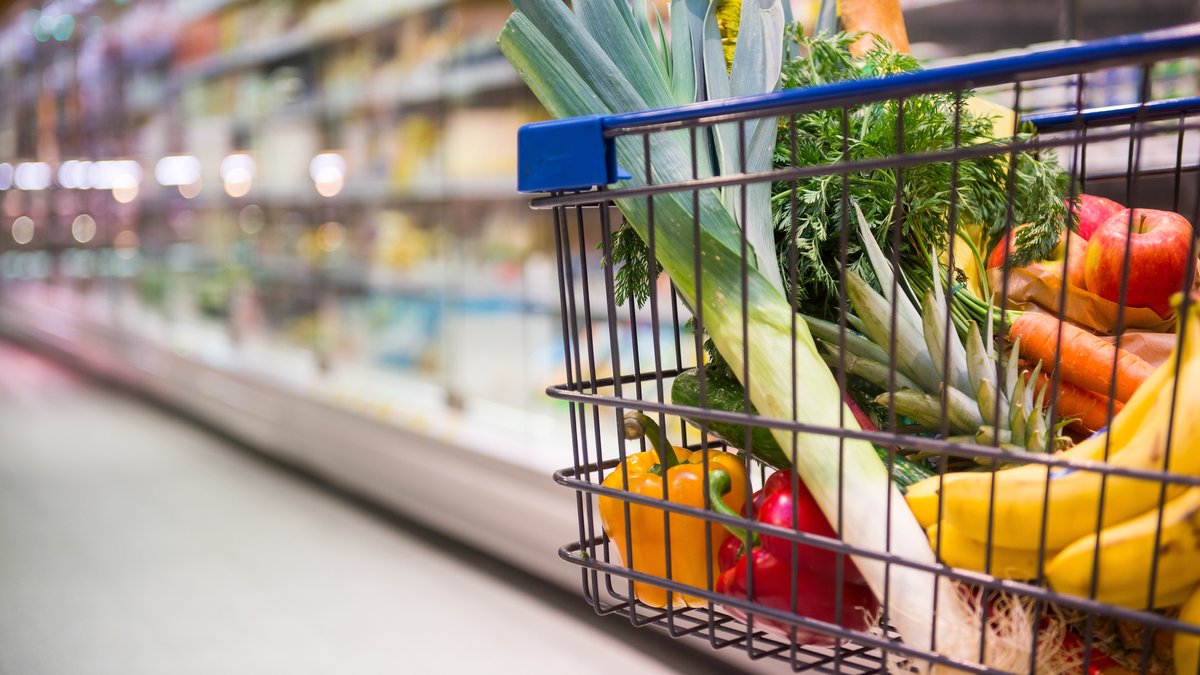 In einem Einkaufswagen liegen Obst und Gemüse. Im Hintergrund sieht man das Kühlregal mit Milchprodukten