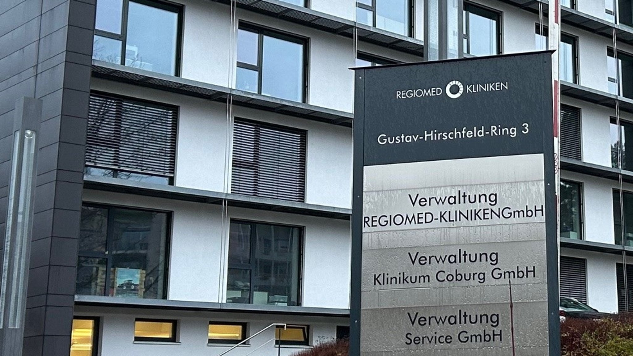 Sitz von Regiomed in Coburg. Zu sehen ist ein Schild "Regiomed Kliniken, Gustav-Hirschfeld-Ring 3; Verwaltung" und im Hintergrund ein Gebäude. 