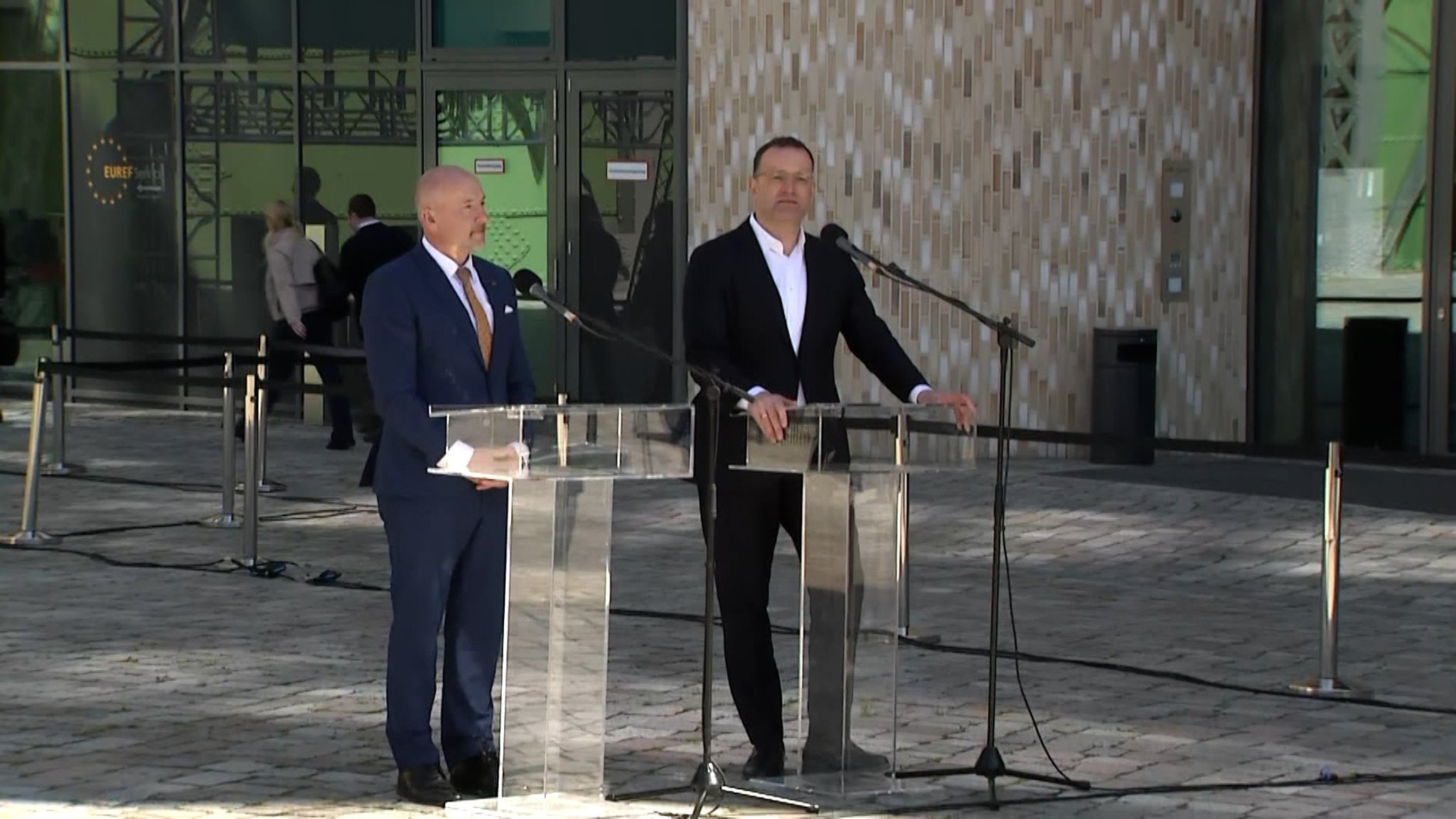 Zwei Männer im Anzug stehen bei einer Pressekonferenz im Freien an Rednerpulten.