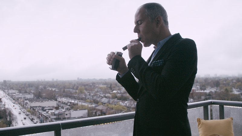 "Push - Für das Grundrecht auf Wohnen"-Filmszene: Mann in Anzug zündet sich auf einer Terrasse eine Zigarre an. 