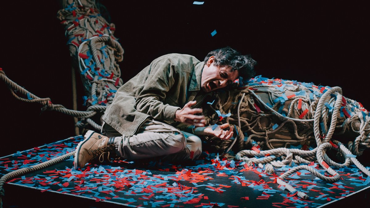 Schauspieler liegt schreiend auf einer Bühne mit bunten Schnipseln, umgeben von Seilen.