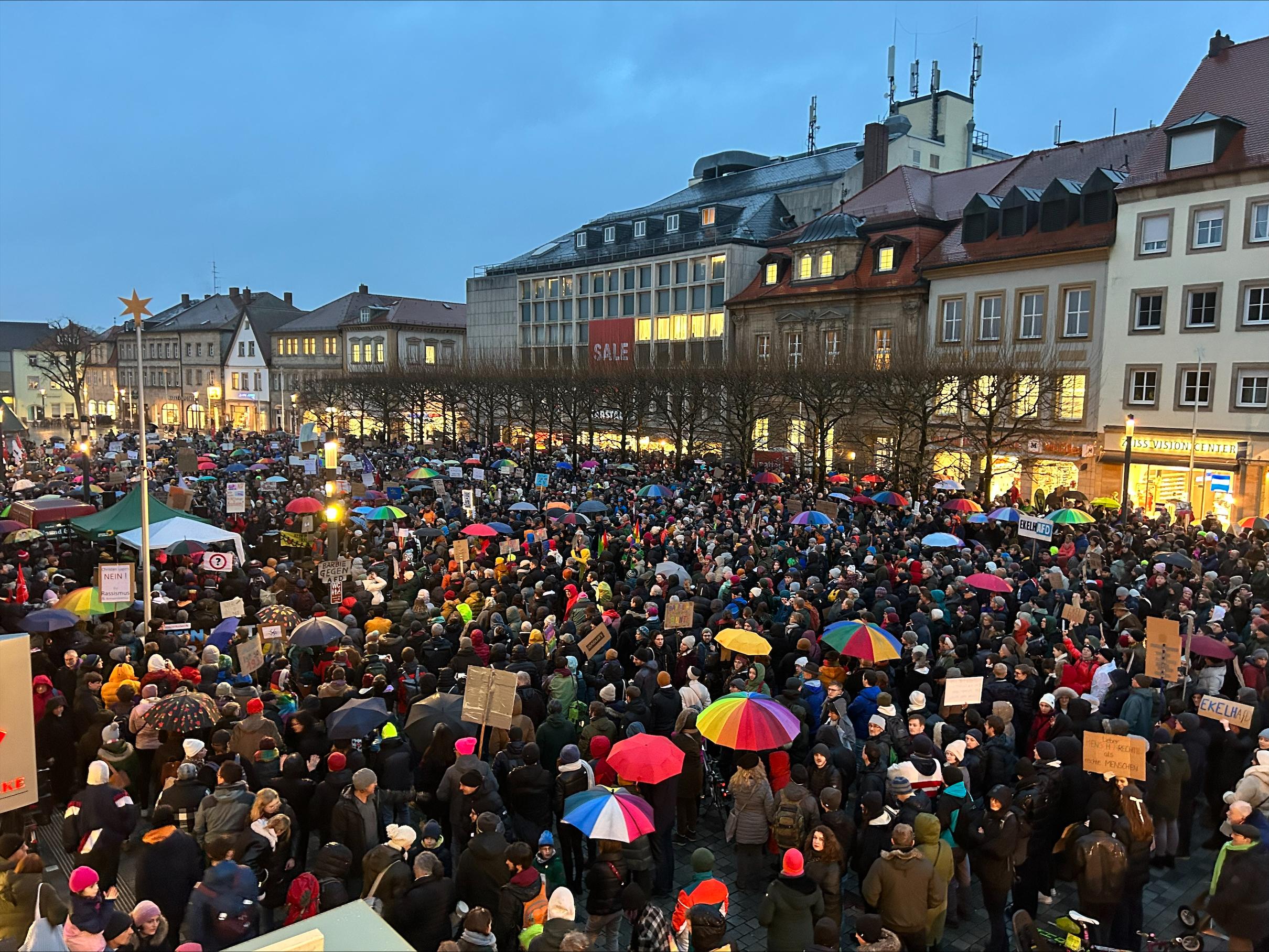 Ein Platz voller Menschen, die bunte Regenschirme halten, betrachtet aus der Vogelperspektive.  