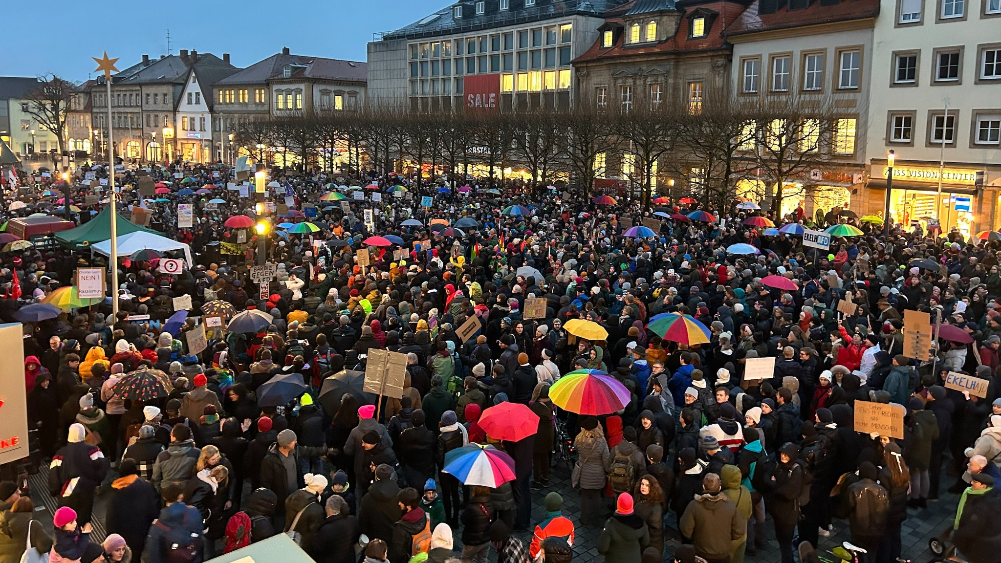 Ein Platz voller Menschen, die bunte Regenschirme halten, betrachtet aus der Vogelperspektive.  