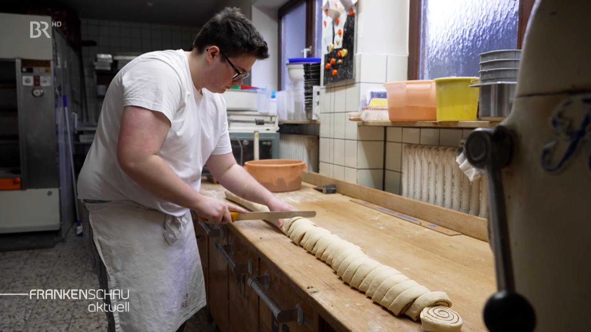 Ein junger Bäcker arbeitet in der Backstube und schneidet eine lange Teigrolle in Scheiben. 