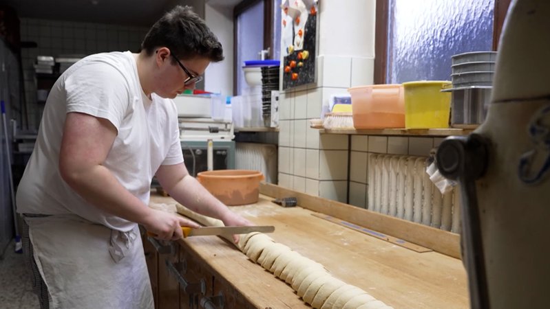 Ein junger Bäcker arbeitet in der Backstube und schneidet eine lange Teigrolle in Scheiben. 