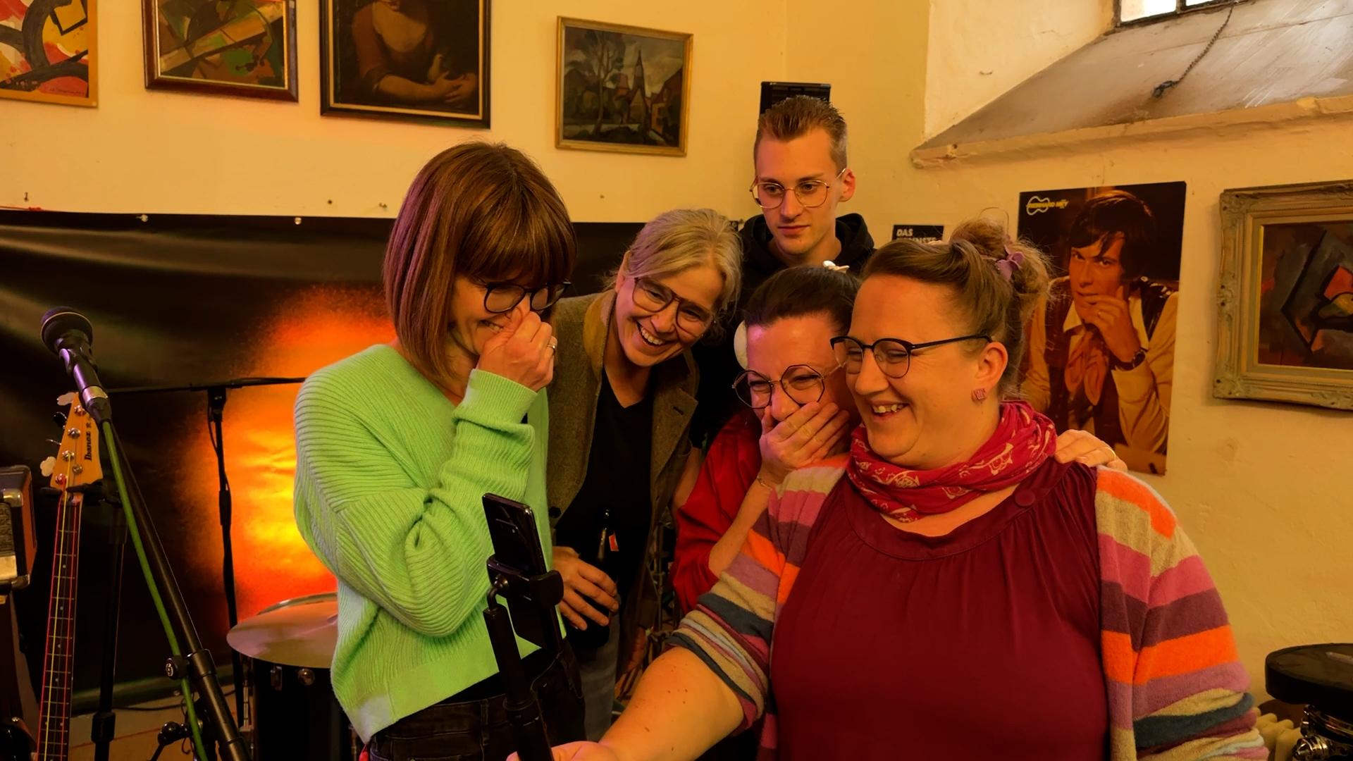 Vier Frauen und ein junger Mann blicken kichernd in ein Handy, mit dem sie sich gerade selbst aufnehmen. 