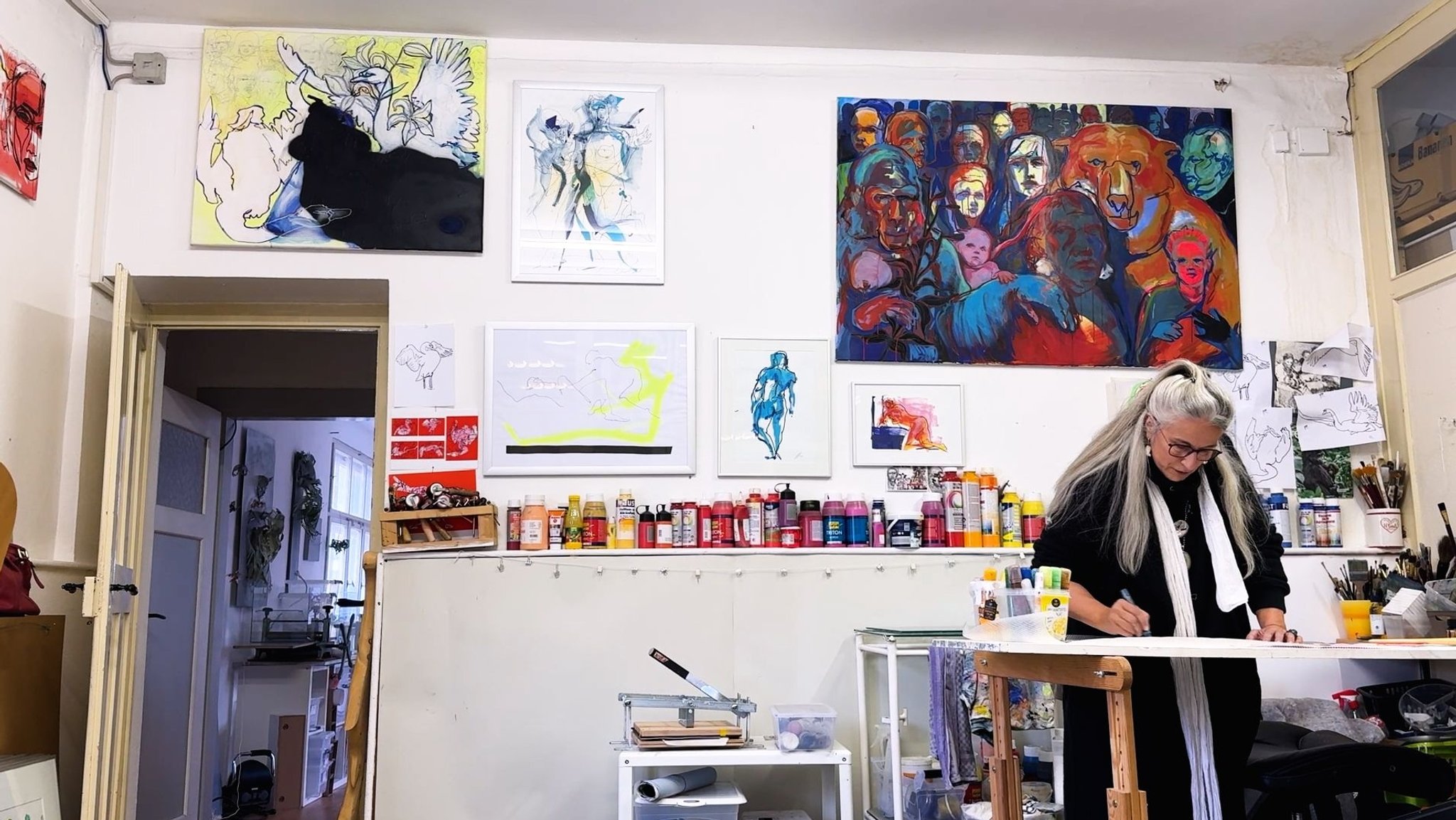 Christiane Gaebert in ihrem Atelier: Sie steht am Tisch und malt, hinter ihr Farbtuben und fertige Bilder. | Bild: BR/ Carolin Hasenauer Christiane Gaebert in ihrem Atelier: Sie steht am Tisch und malt, hinter ihr Farbtuben und fertige Bilder.