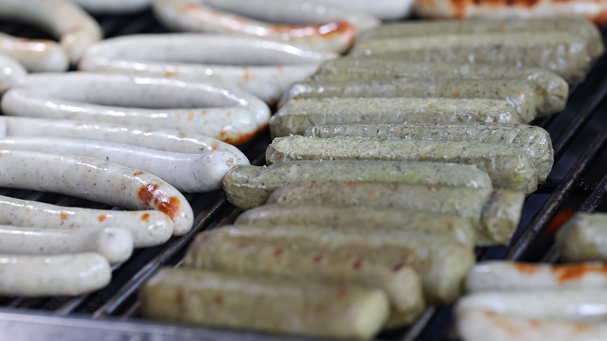 Echte Bratwürste (l) und vegane Bratwürste (r) liegen nebeneinander auf dem Holzkohlengrill. 