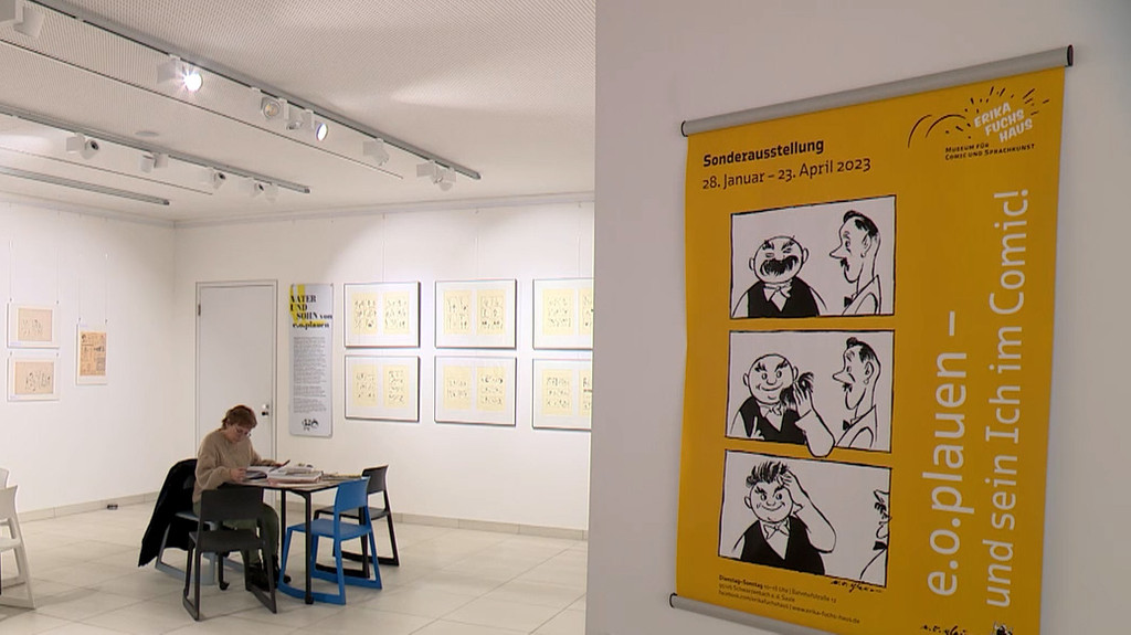 In einem Ausstellungsraum sitzt eine Frau an einem Tisch und liest, an der Wand hängen Zeichnungen und ein gelbes Plakat mit der der Aufschrift "e.o.plauen und sein Ich im Comic".