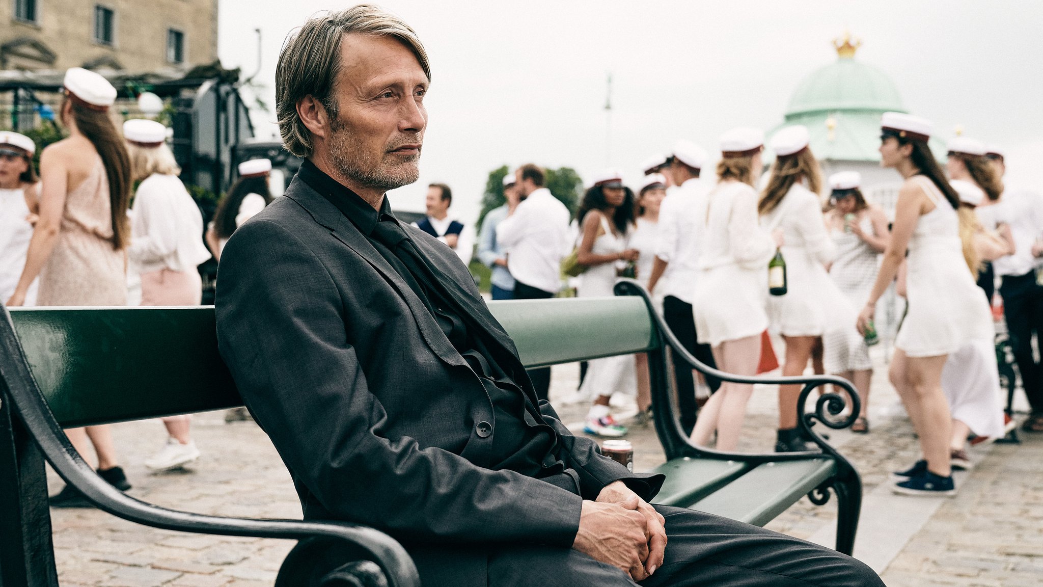 Mads Mikkelsen sitzt ganz allein auf einer Bank, während neben ihm das Leben tobt.