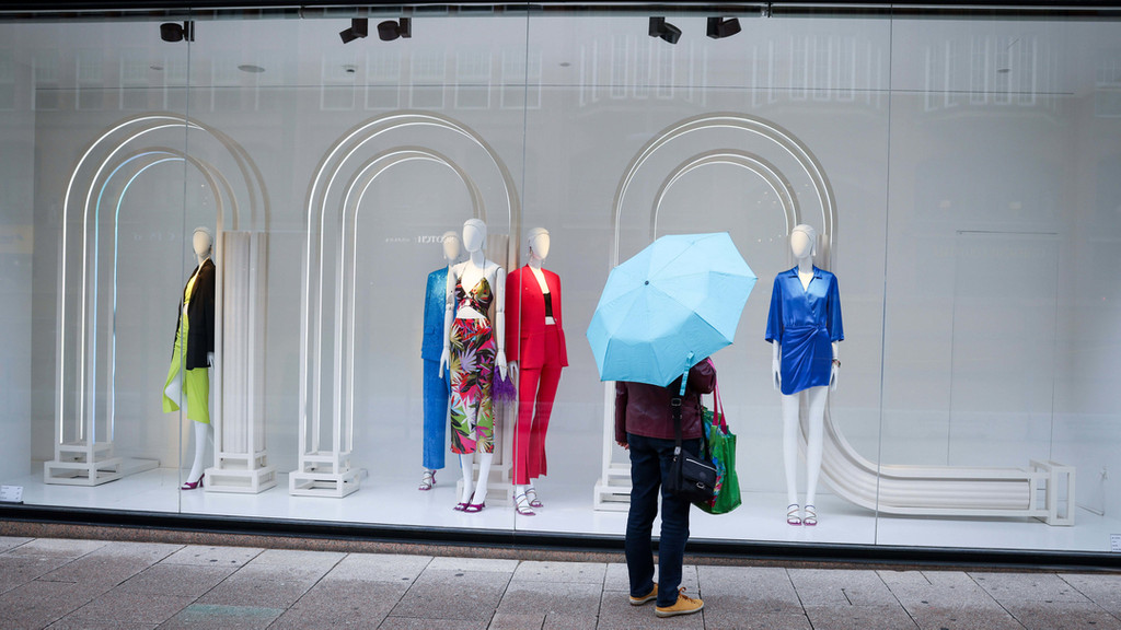 Eine Passantin mit Regenschirm steht in der Innenstadt vor einem Schaufenster mit Sommerkleidung.