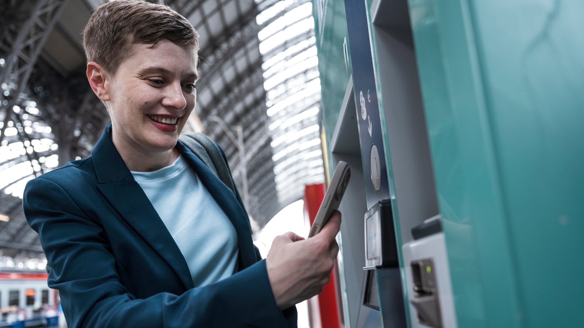 (Symbolbild) Eine Frau hält ihr Smartphone an einen Bahnautomaten. 