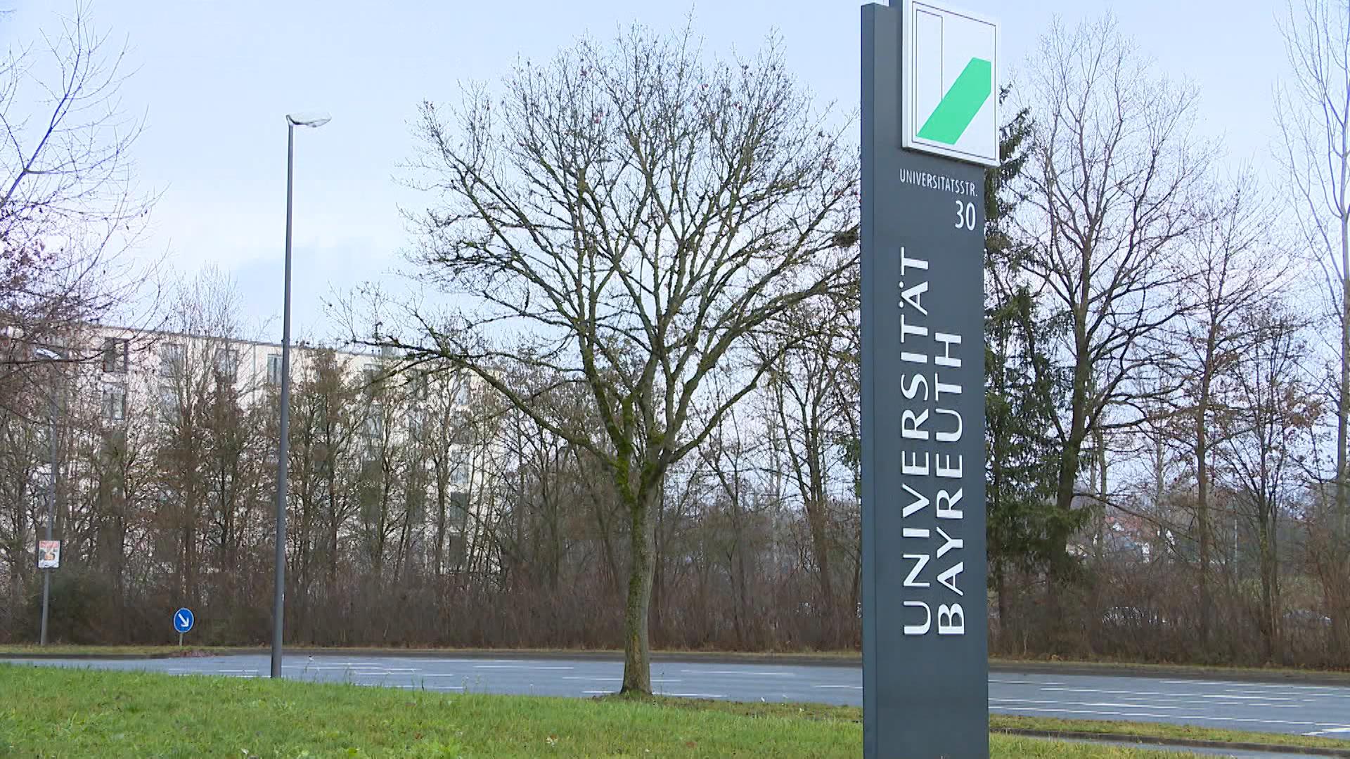 Blick auf ein Gebäude, davor eine Tafel mit der Aufschrift "Universität Bayreuth". 