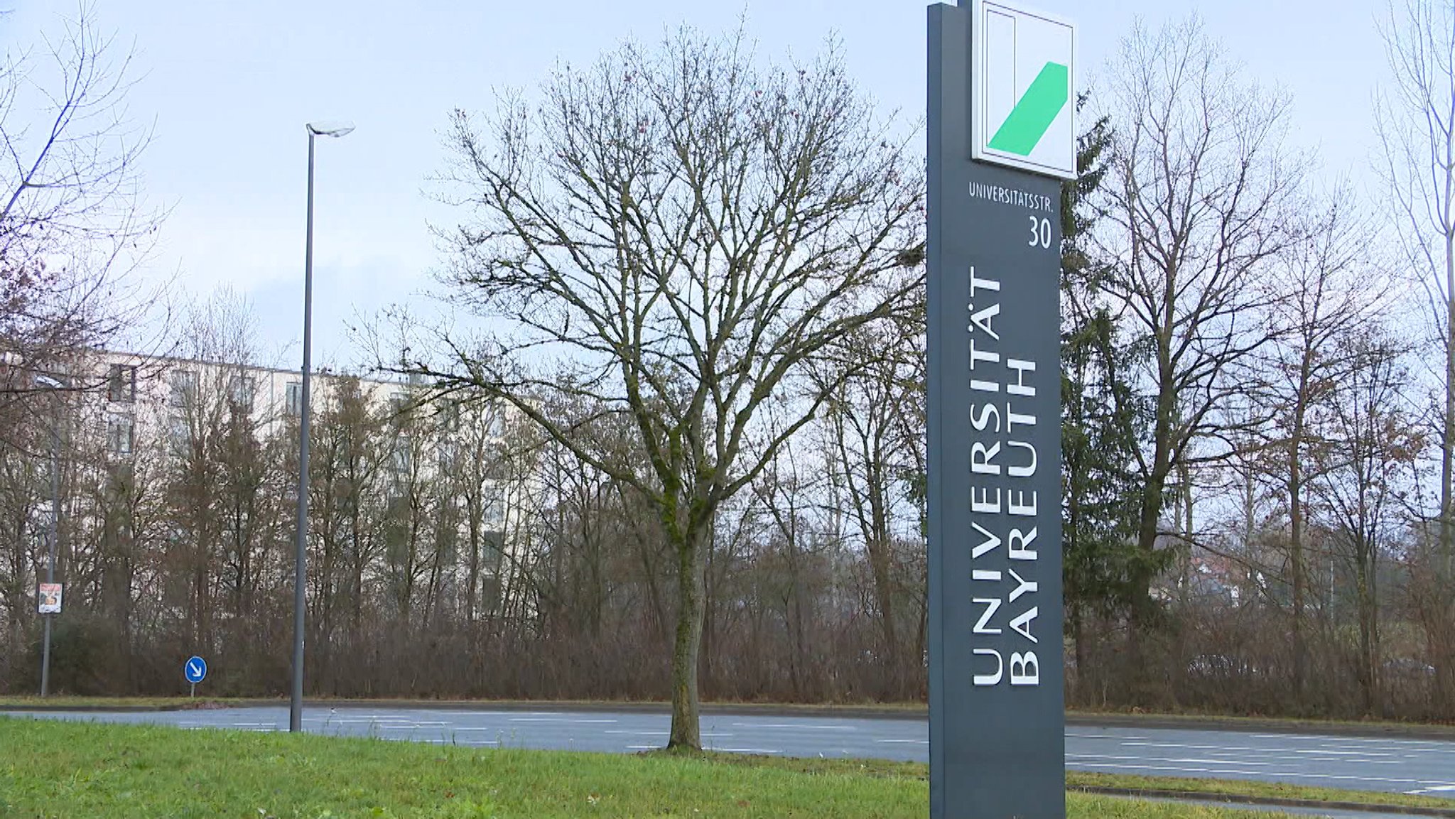 Blick auf ein Gebäude, davor eine Tafel mit der Aufschrift "Universität Bayreuth". 