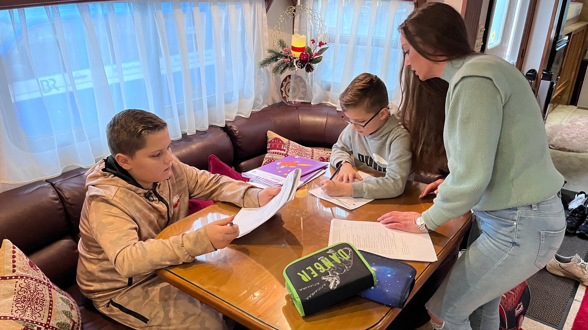 Zwei Kinder sitzen an einem Tisch und machen Hausaufgaben. Ihre Mutter steht am Tisch und spricht mit ihnen.