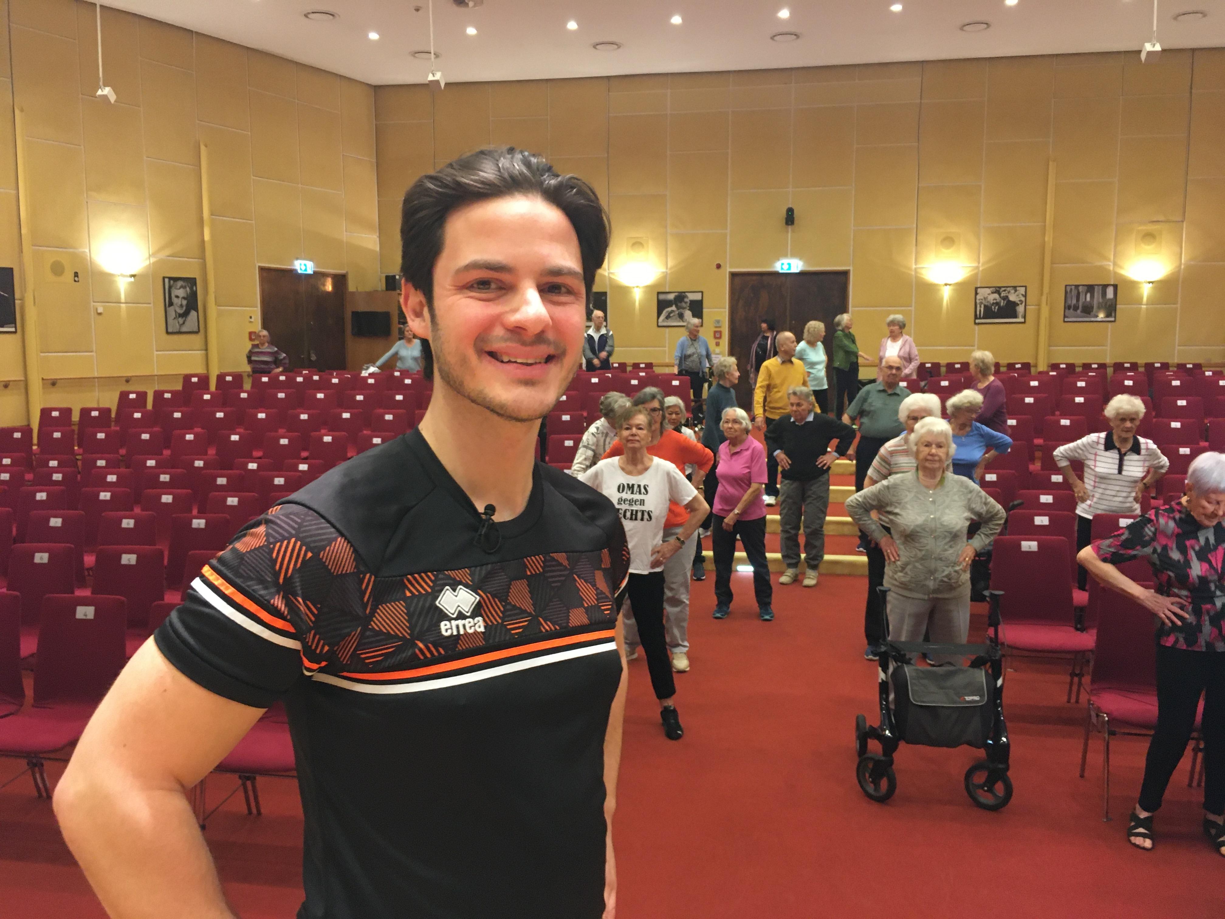 Fitnesscoach Marco del Popolo, im Hintergrund Seniorinnen und Senioren in einem Saal mit Stuhlreihen