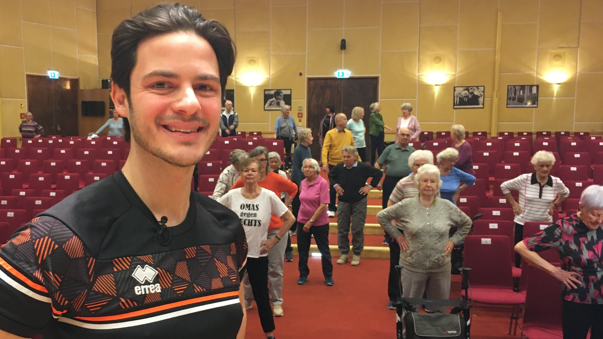 Fitnesscoach Marco del Popolo, im Hintergrund Seniorinnen und Senioren in einem Saal mit Stuhlreihen