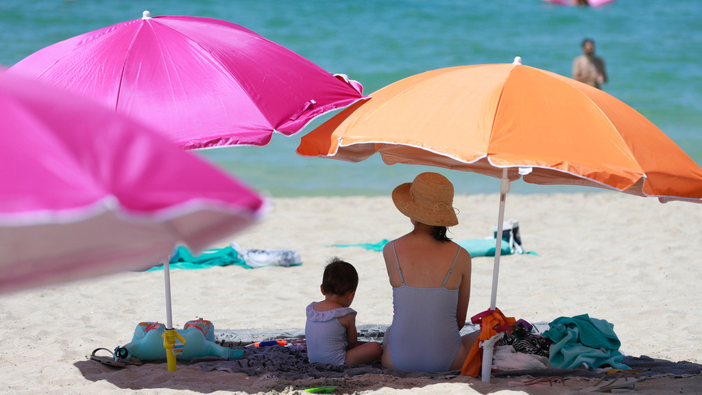 Eine Frau sitzt neben ihrem Kind unter einem Sonnenschirm an einem warmen Sommertag am Strand.