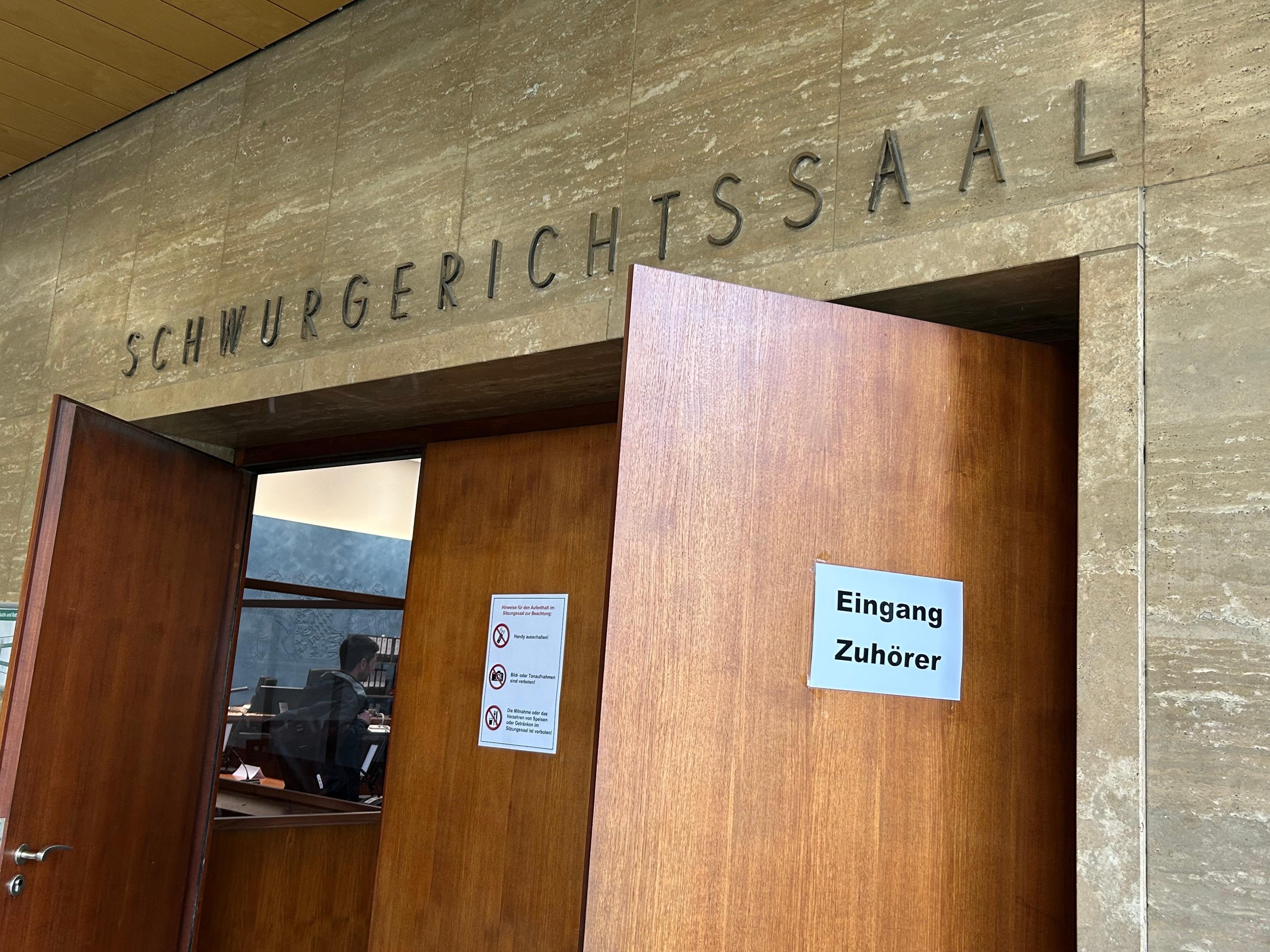 "Schwurgerichtssaal" steht über einer Holztüre, die offen steht. Im Gerichtsaal ist eine Person zu erkennen. 