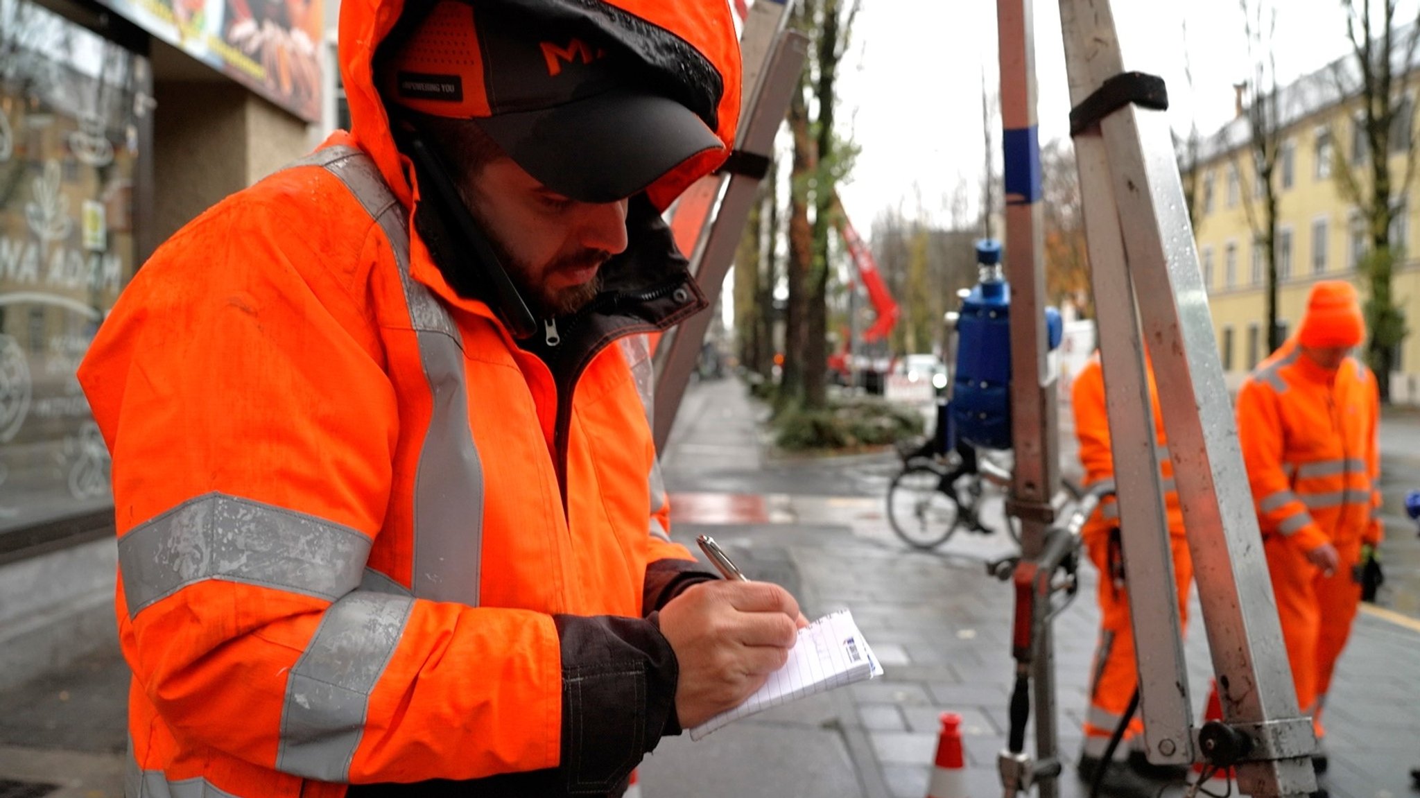 Foto: Ein Mann in Arbeitskleidung steht an der Straße, telefoniert und macht sich Notizen in seinen Block.