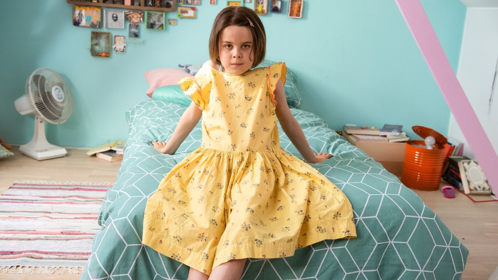 Oskar alias Lili sitzt in ihrem gelben Sommerkleid auf dem Bett im Kinderzimmer. Wand und Bett sind lindgrün.
