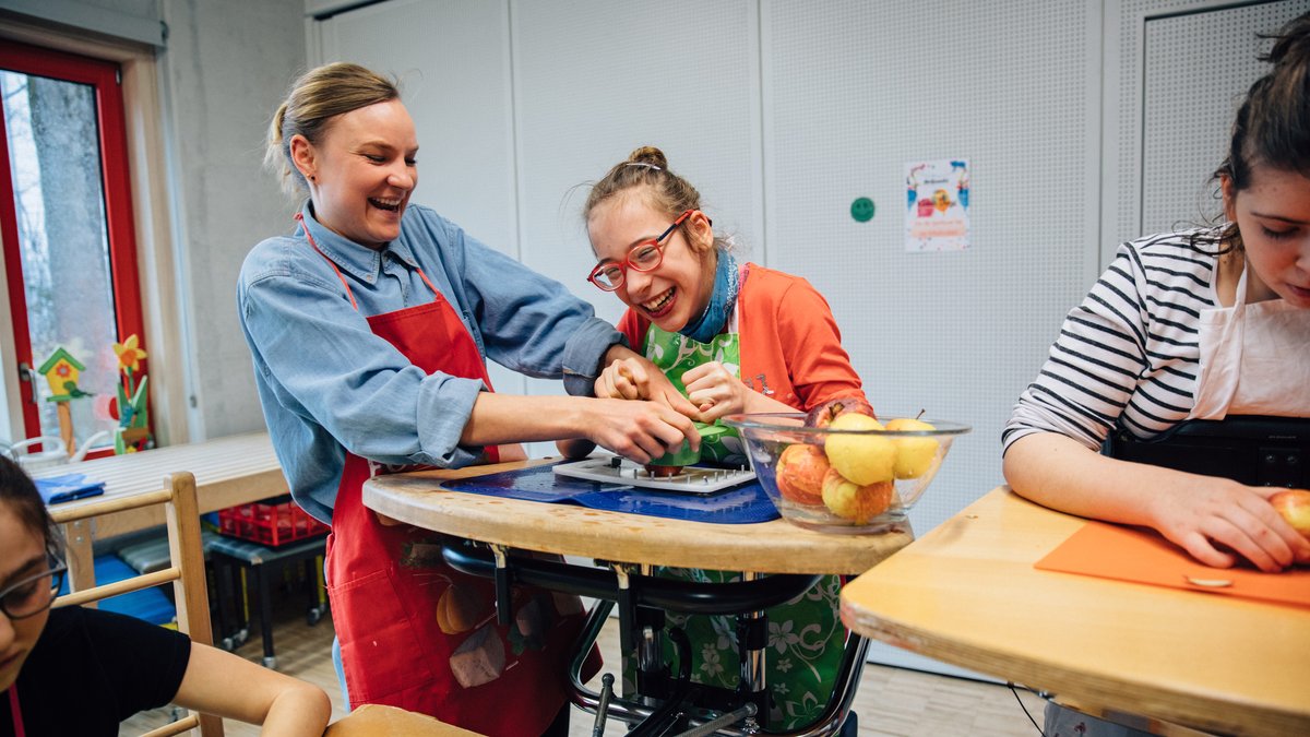 Eine Frau in Schürze, die einem jungen Mädchen mit Behinderung beim Kochen hilft