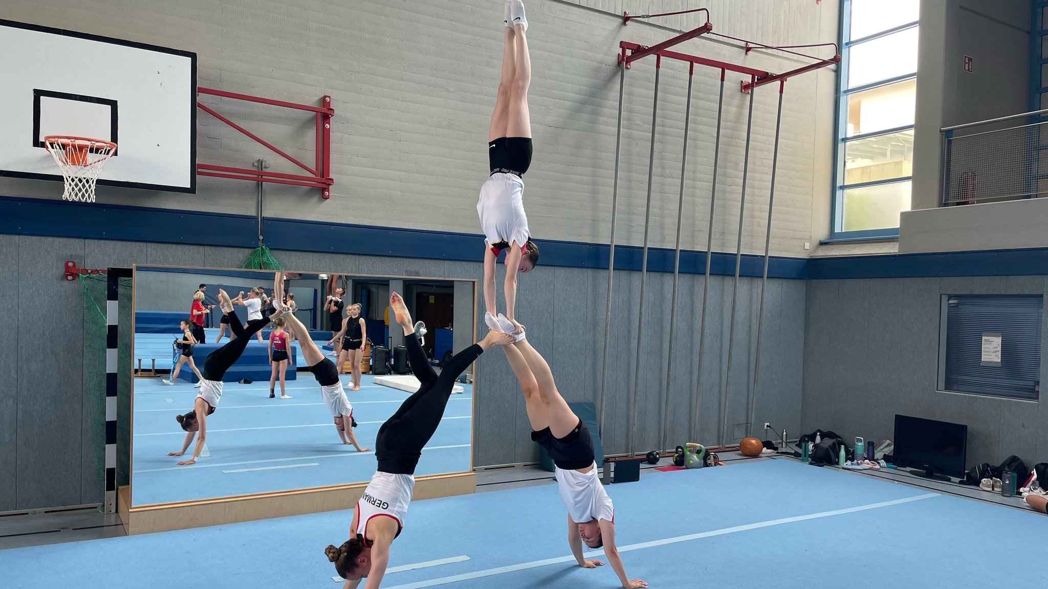 Drei Frauen stehen in einer Handstandpyramide in einer Turnhalle vor einem großen Spiegel.