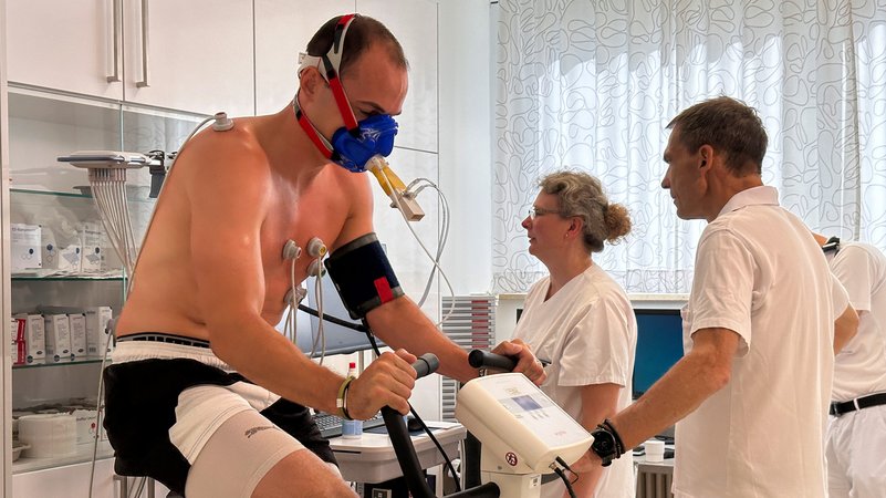 Mann auf einem Ergometer, zwei Ärzte im Hintergrund. | Bild: BR/Henry Lai Mann auf einem Ergometer, zwei Ärzte im Hintergrund.