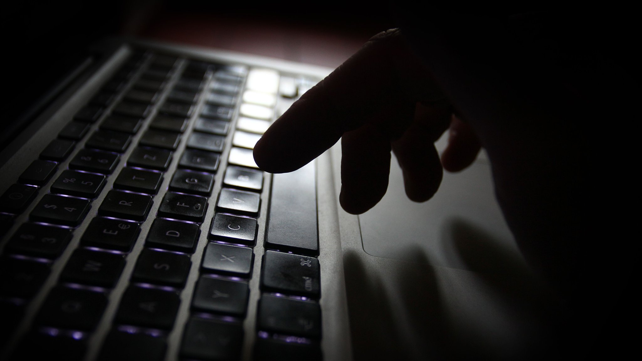 Man sieht eine Hand, die über einer Laptop-Tastatur schwebt. Das Bild ist insgesamt dunkel gehalten.
