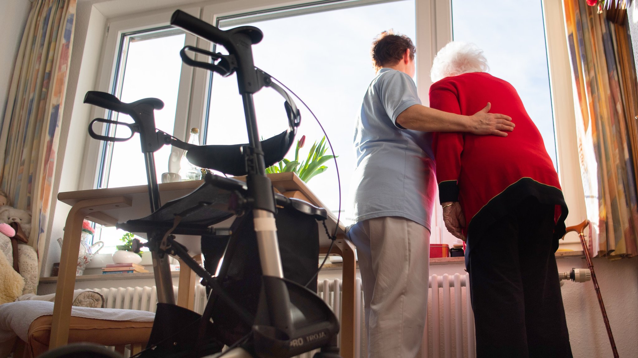 Eine Pflegekraft steht mit der Bewohnerin eines Pflegeheims am Fenster und schaut hinaus, im Vordergrund steht ein Rollator.