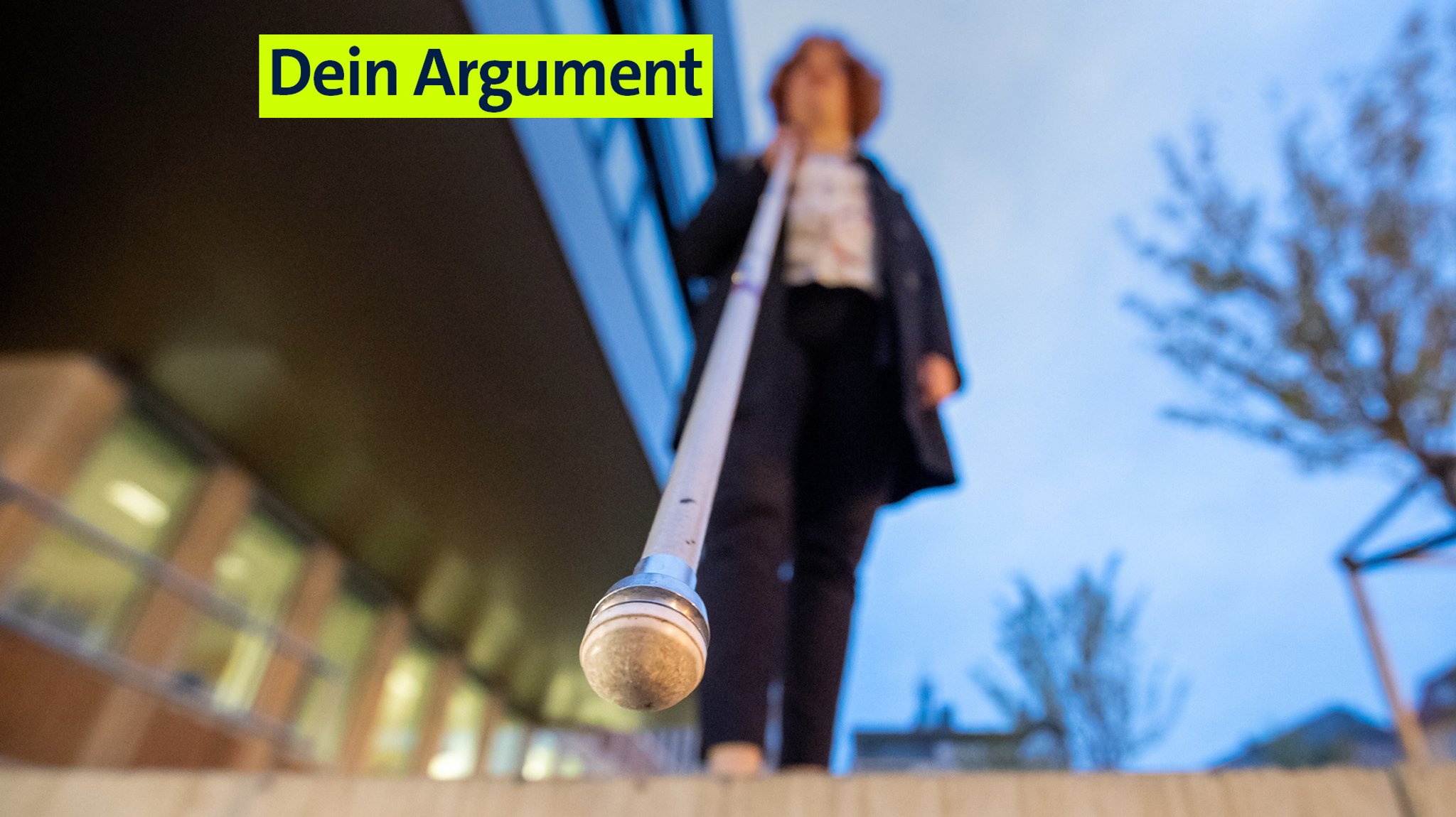 Eine Frau geht am Abend mit einem Blindenstock auf Treppenstufen zu. Im Hintergrund sind rechts zwei Bäume zu sehen, links steht ein Gebäude. 