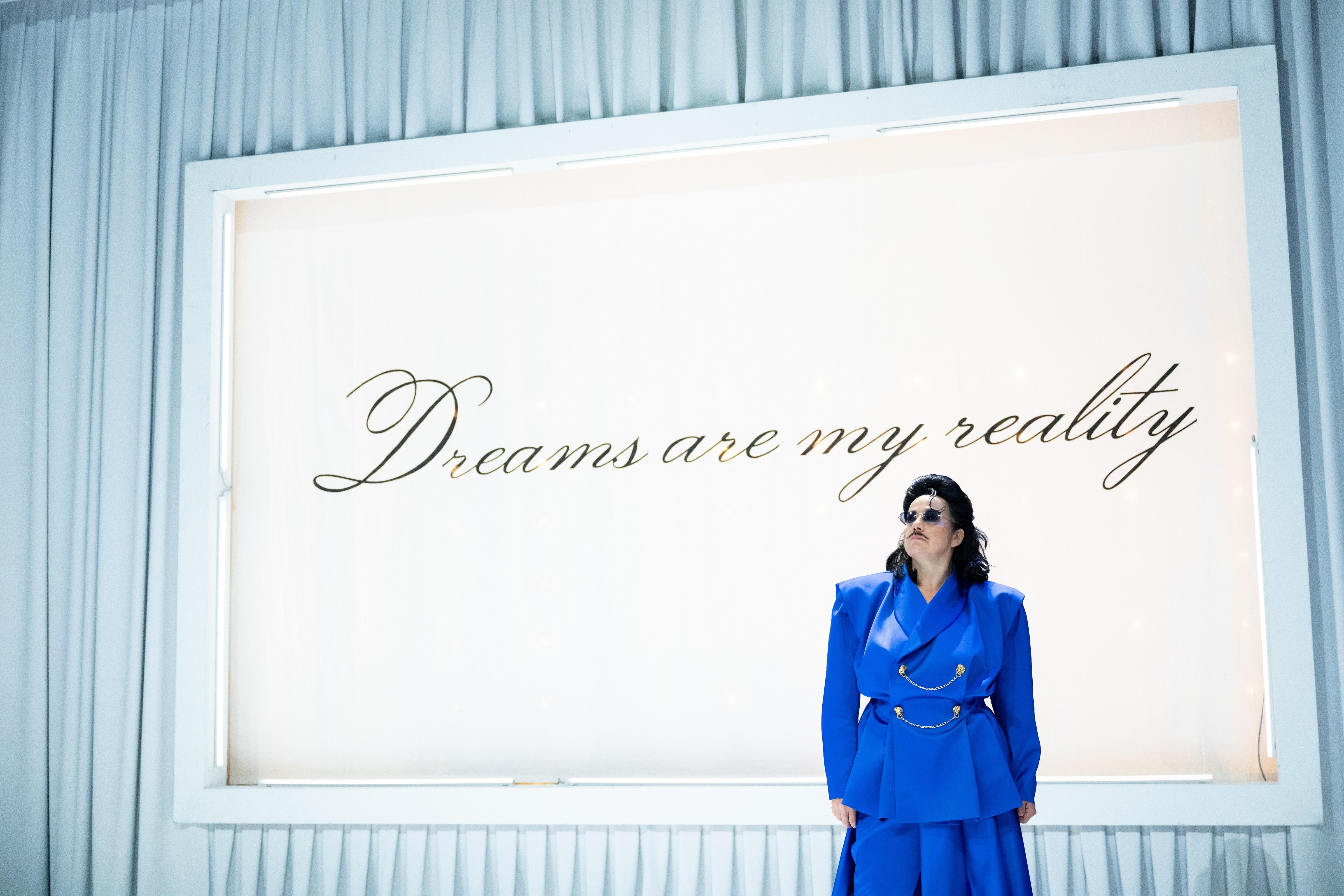 Figur mit knallblauem Jackett, schwarzen Haaren und Sonnenbrille vor weißer Wand auf der in Schreibschrift steht: "Dreams are my reality"