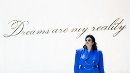 Figur mit knallblauem Jackett, schwarzen Haaren und Sonnenbrille vor weißer Wand auf der in Schreibschrift steht: "Dreams are my reality" | Bild:© Sandra Then
