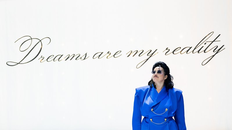 Figur mit knallblauem Jackett, schwarzen Haaren und Sonnenbrille vor weißer Wand auf der in Schreibschrift steht: "Dreams are my reality"