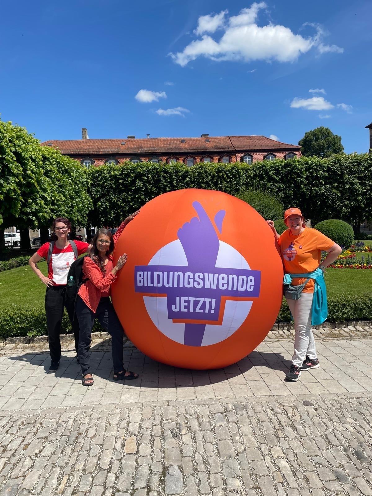 Drei Frauen stehen an einem orangenen Ball, auf dem "Bildungswende Jetzt!" steht.