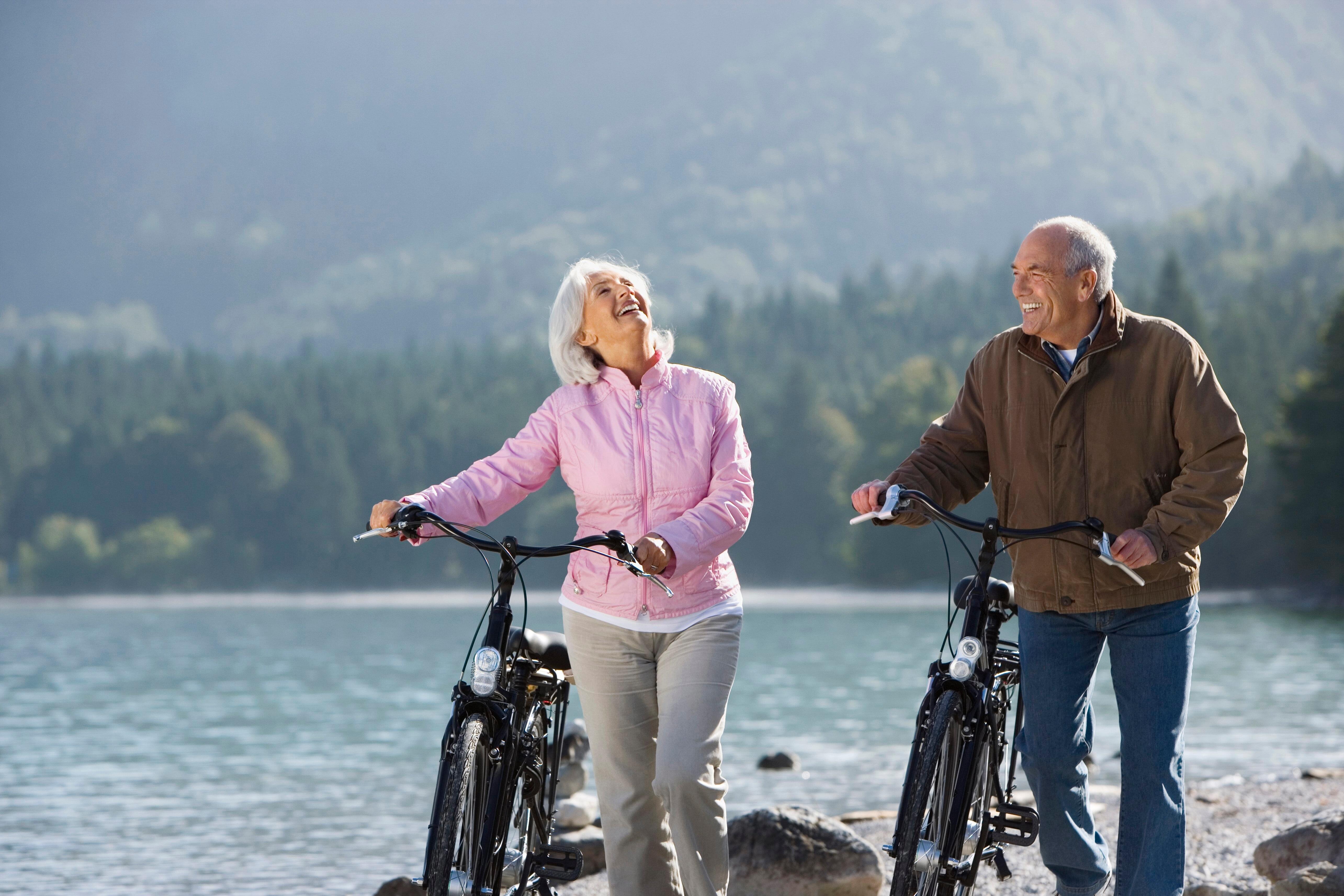 (Symbolbild) Zwei ältere Menschen mit Fahrrädern vor einer See-Kulisse