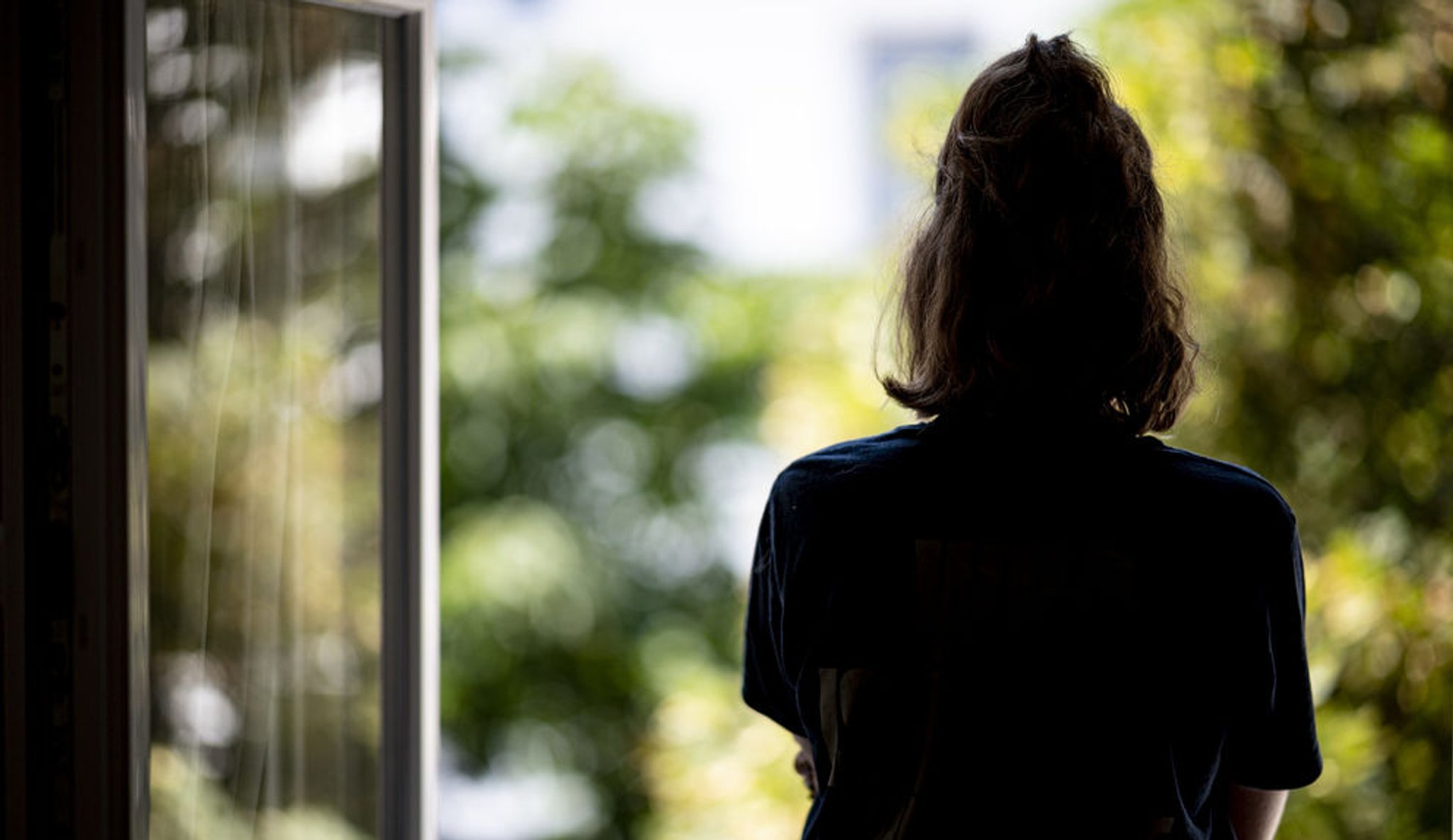 (Symbolbild) Eine Frau steht an einem Fenster und sieht heraus, man sieht sie nur von hinten.