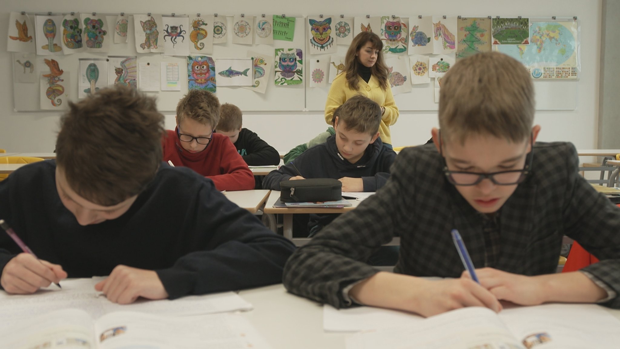 In einer Schulklasse sitzen Jugendliche beim Schreiben eines Texts am Schreibtisch. Im Hintergrund steht die Lehrerin der Brückenklasse an der Realschule Freiham. 